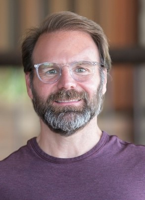 close-up of man with brown hair and beard, glasses, wearing a maroon t-shirt